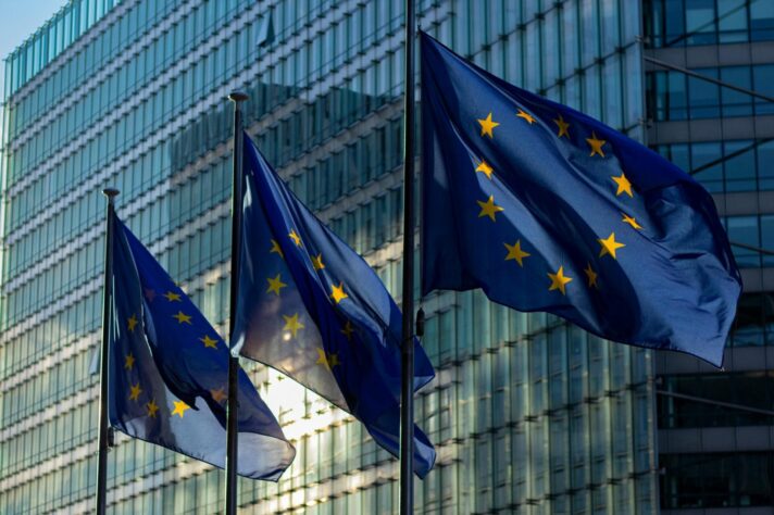 Image shows three EU flags flying low in front of a modern sky-rise office block. The sun is setting.