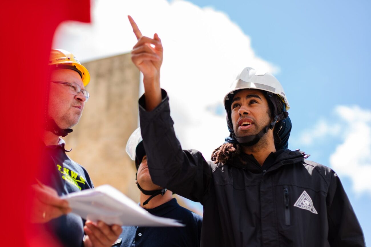 A group of construction workers wearing helmets review documents outdoors as one worker gestures upwards, pointing toward something off frame against a bright sky.