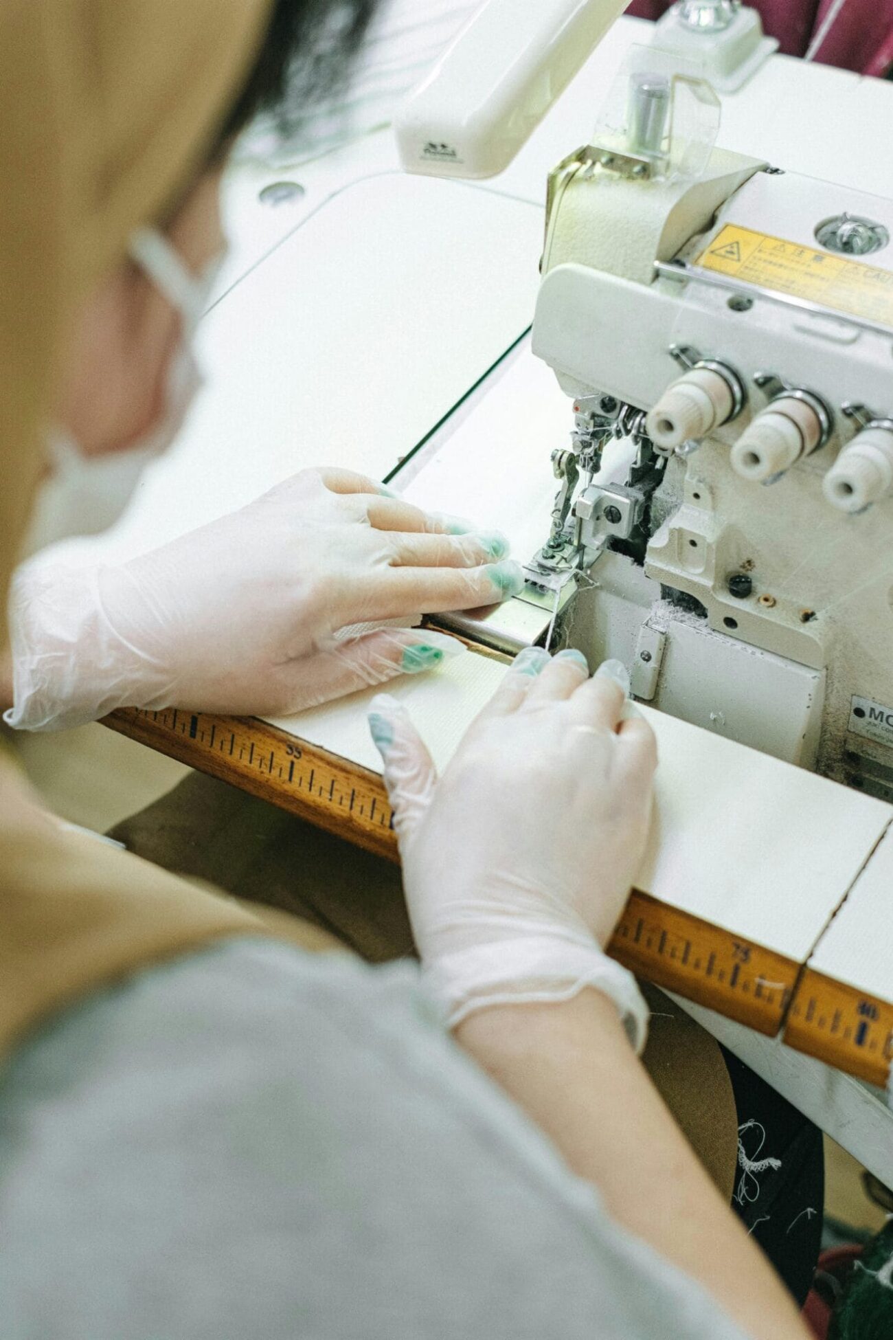 A person wearing disposable gloves operates a sewing machine, guiding fabric through the needle mechanism.