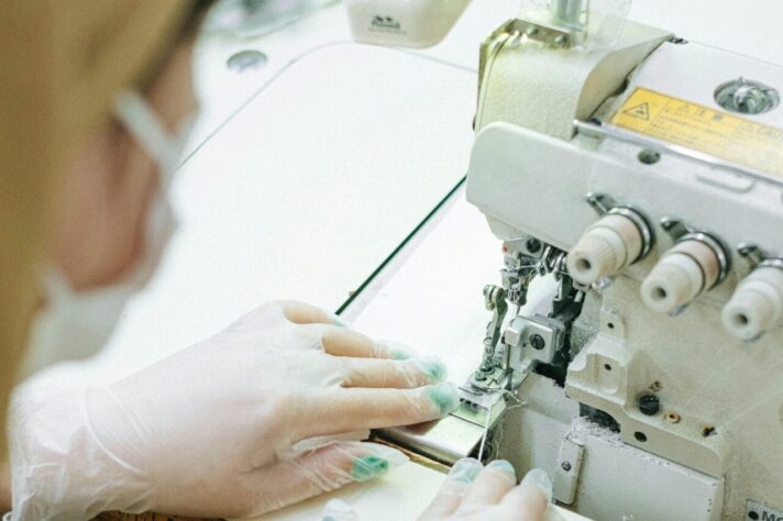 A person wearing disposable gloves operates a sewing machine, guiding fabric through the needle mechanism.