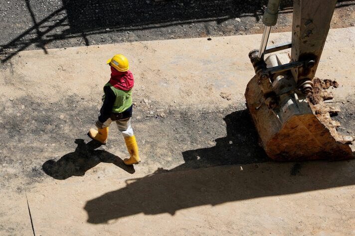 Construction worker walks across the yard. He is wearing yelow wellies, a hard ht and gloves.