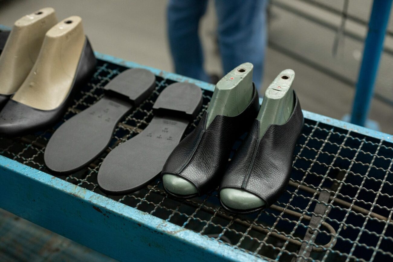 Black leather shoe uppers fitted onto green and beige moulds sit on a metal conveyor alongside separate rubber soles in a footwear manufacturing facility.