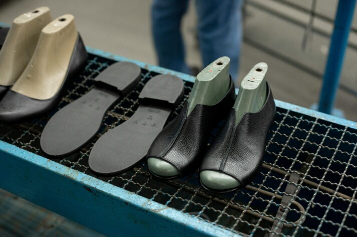 Black leather shoe uppers fitted onto green and beige moulds sit on a metal conveyor alongside separate rubber soles in a footwear manufacturing facility.