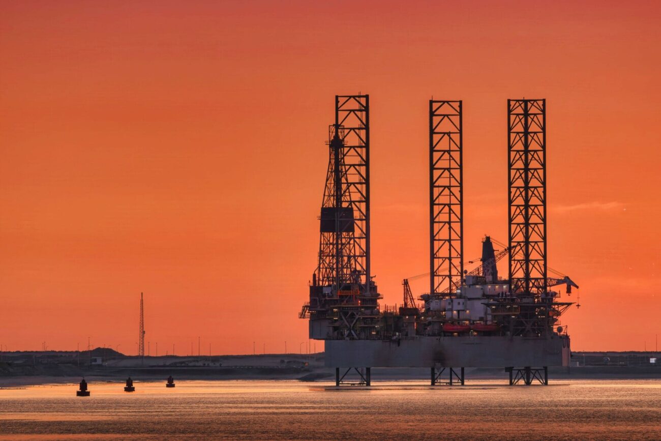Silhouette of an offshore oil drilling platform with tall lattice towers standing in calm water at sunset, set against a vivid orange sky; small buoys and a low shoreline are visible in the foreground and background.