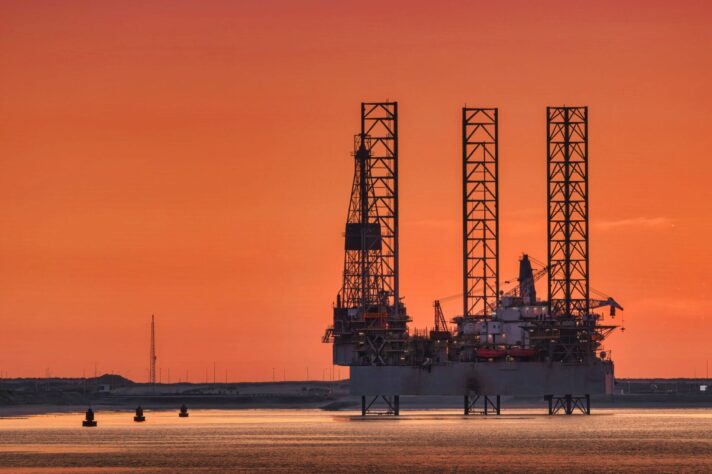 Silhouette of an offshore oil drilling platform with tall lattice towers standing in calm water at sunset, set against a vivid orange sky; small buoys and a low shoreline are visible in the foreground and background.